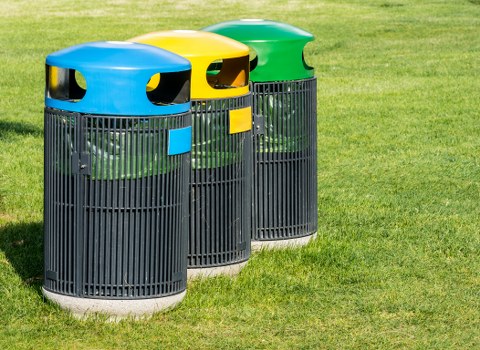 Three outdoor litter bins positioned on a grassy area, each with a cylindrical black plastic body featuring vertical slats, and distinctively colored domed lids in blue, yellow, and green. The lids have openings for waste disposal and match in color with small rectangular signs on the front of each bin, indicating separate waste or recycling categories. The bins are situated in a neat row, with the blue-lidded bin closest to the viewer, followed by the yellow and green-lidded bins, which are slightly behind and to the right. The background consists of well-maintained green grass, suggesting a garden, park, or communal outdoor space in Hoxton, London, where Garden Clearance Hoxton operates. The lighting appears natural, illuminating the bins evenly without harsh shadows, emphasizing cleanliness and organization. This image depicts typical rubbish disposal points for waste collection, relevant to residential or community-based rubbish removal services in London.