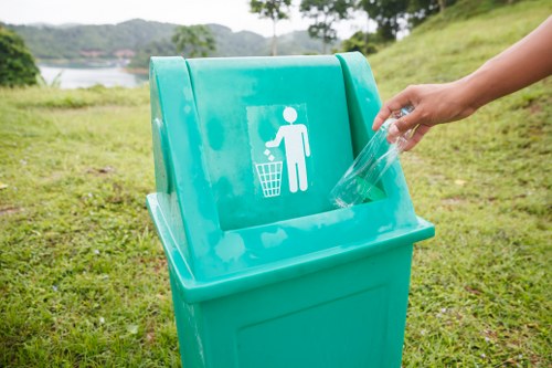 A turquoise plastic rubbish bin situated outdoors on a grassy area with a landscape view of trees and water in the background. The bin has a white printed symbol of a person disposing of waste into a bin, indicating it's intended for general refuse. A person's hand is seen placing a transparent plastic bag into the bin, which is positioned on a flat surface of short, green grass. The scene appears to be in a park or open space, possibly in or near Hoxton, with natural daylight illuminating the environment. The image reflects a focus on proper waste disposal, relevant to garden clearance and rubbish removal services conducted by Garden Clearance Hoxton in the local area, suggesting environmentally conscious waste management practices.