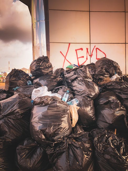 A close-up view of a collection of mixed waste materials piled on the ground outdoors. The trash includes several black plastic garbage bags, some of which are torn or partially open, revealing assort