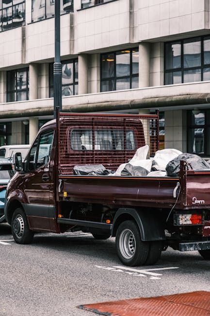 A small, dark red pickup truck parked on an urban street in front of a modern building with large glass windows and a concrete facade. The truck's flatbed is loaded with a variety of discarded waste m