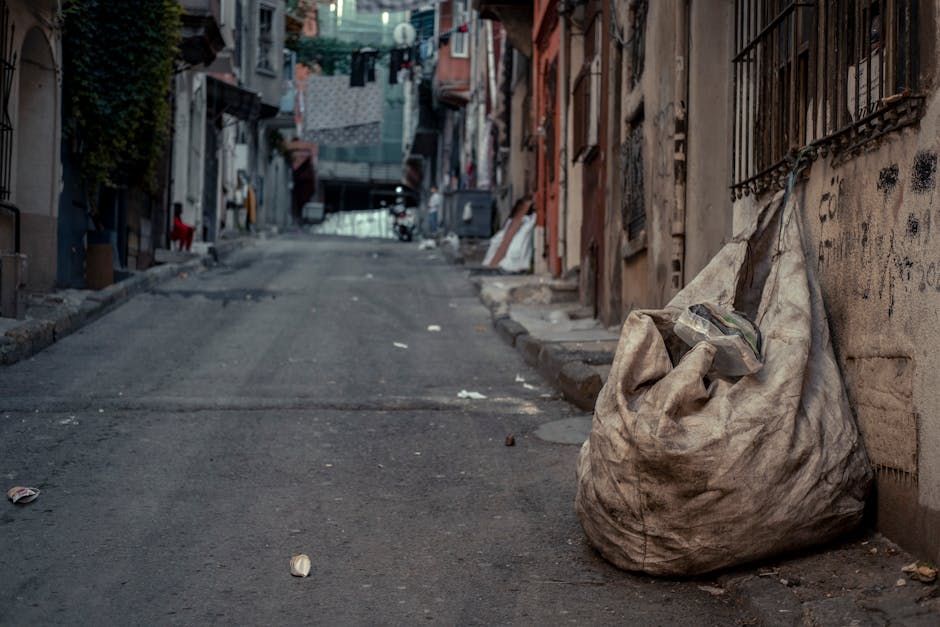 An alleyway with a narrow passage between two urban buildings, cluttered with discarded rubbish, including a large black trash bag in the foreground and stacked cardboard boxes and plastic waste piled