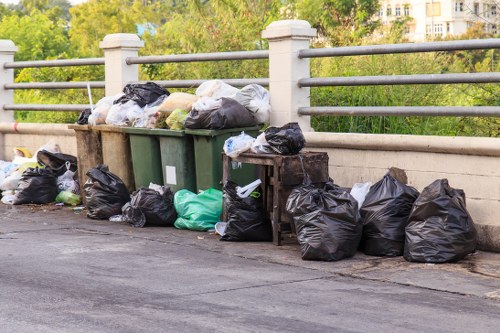 A collection of black and green refuse bags, along with a few white and transparent bags, are piled along the edge of a paved urban street beneath a simple metal railing supported by concrete posts. Some bags appear to contain household waste, while others may hold garden or miscellaneous rubbish. The bags are positioned in front of and partially leaning against a row of three large wheelie bins, with one green bin visible among them. The background features a green landscape with trees and a hint of residential buildings, suggesting a quiet local area possibly within or near Hoxton. The scene is lit by natural daylight, illuminating the varied textures of the plastic bags and the smooth surfaces of the bins, indicative of typical rubbish removal activity managed by local waste collection services like Garden Clearance Hoxton. This image visually captures the sort of rubbish accumulation that requires professional clearance to ensure tidy and environmentally responsible disposal in the local community.